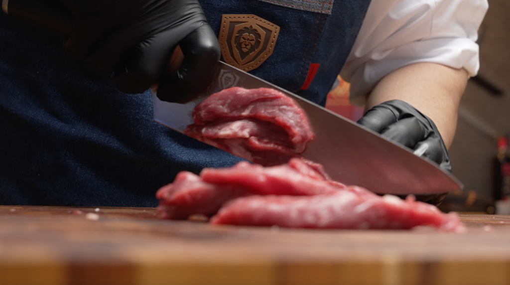 A man's hand with gloves slicing meat using the Vanquish Series Chef's Knife 8"