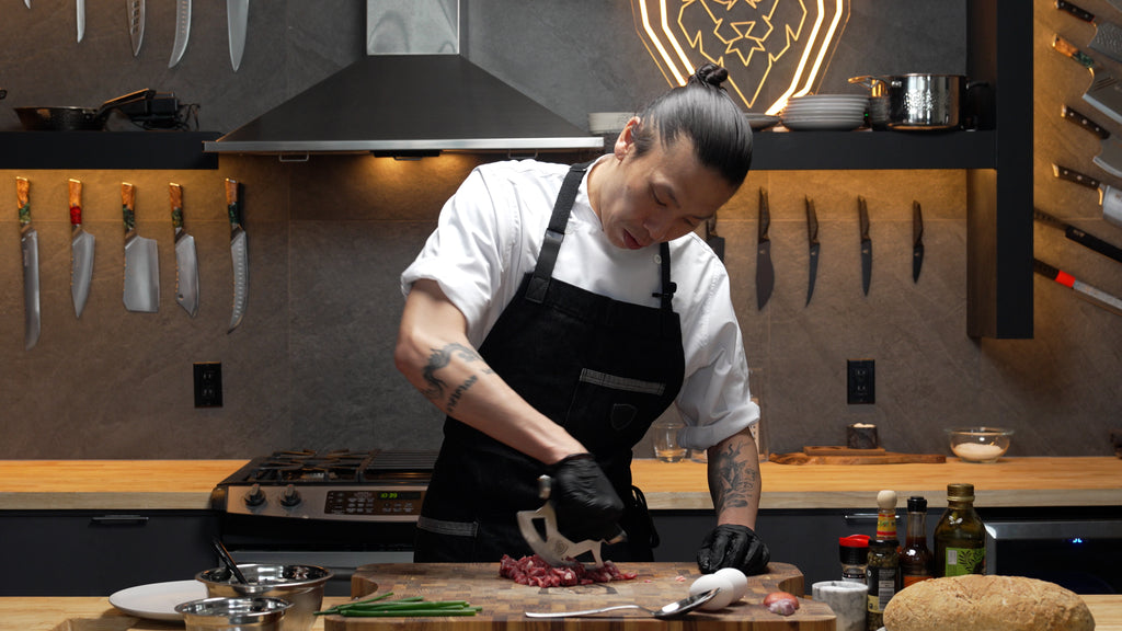 A man with black apron and gloves chopping meat using the Gladiator Series Ulu Rocking Knife 7"